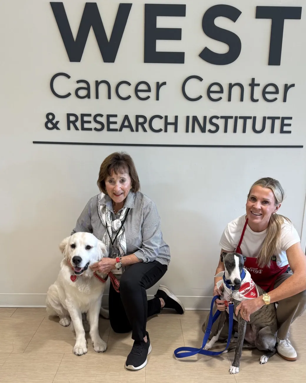 Jo Anne Fusco and Cassie Mayatte, each kneeling beside a therapy dog, under the West Cancer Center & Research Institute sign